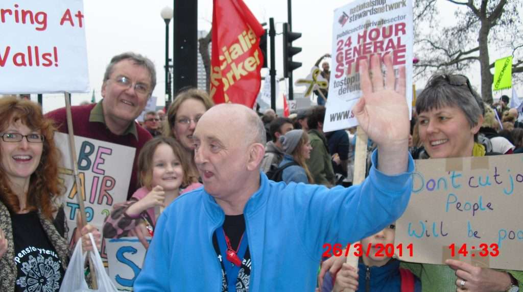 A photo of Brian March in a blue jacket. He is at the front of a group of people who are holding placards.