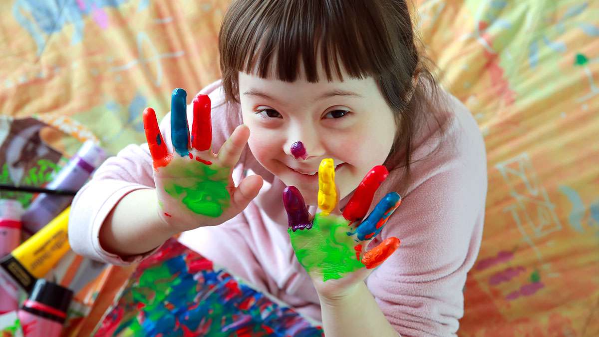 A child with addition support needs is smiling at the camera and holding up painted hands.