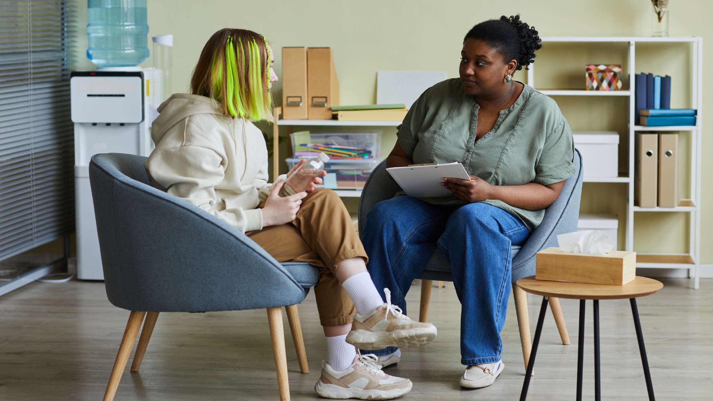 Two young women sat facing each other. One has a clipboard and is listening. The other is talking and holding a water bottle.