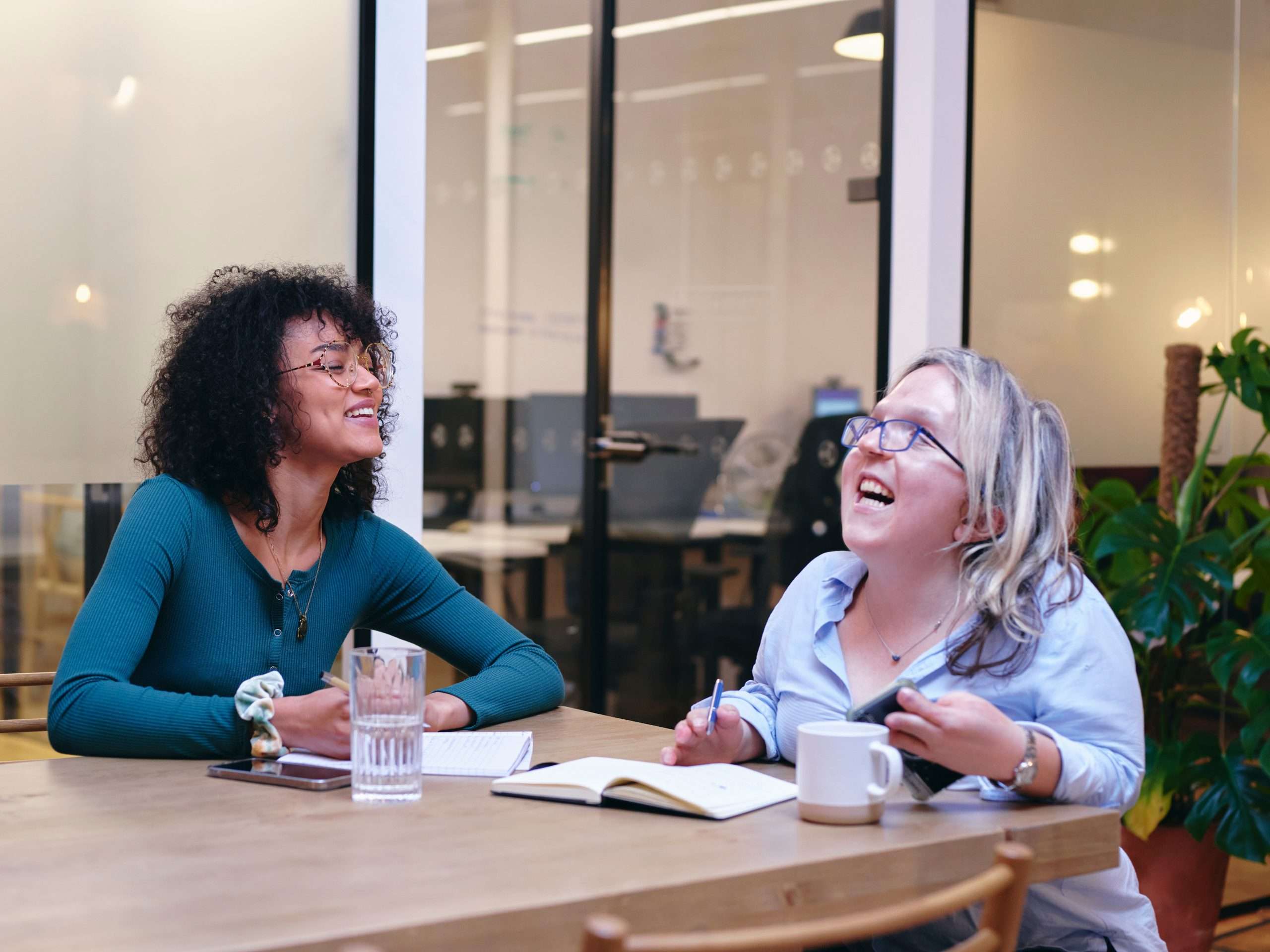 Two people sitting at a table. They have note books, drinks and are laughing. One may be disabled.