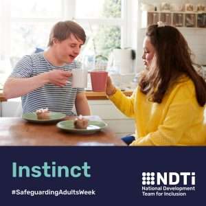 Two people are sat at a table with coffee mugs and cake. They are chatting and smiling. One may have a learning disability.
