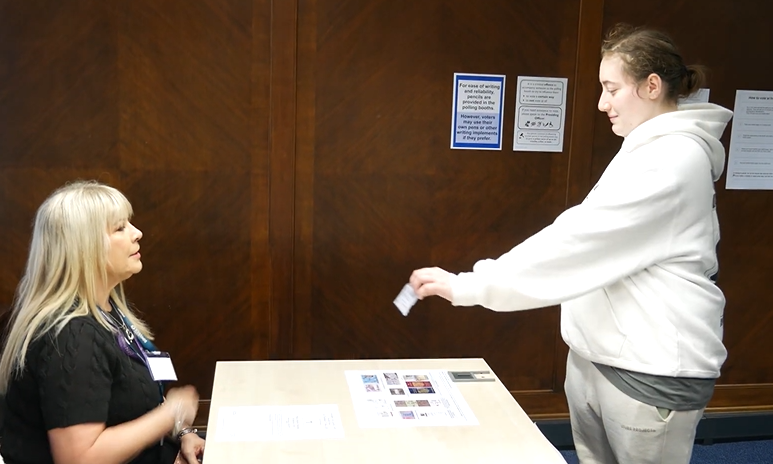 A person is standing at a desk. They are showing the person behind the desk their photo ID.