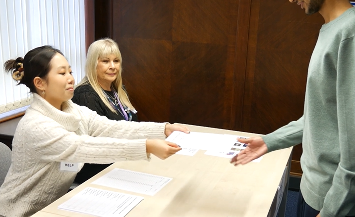 Two people behind a desk handing a piece of paper to a person standing on the other side of the desk.