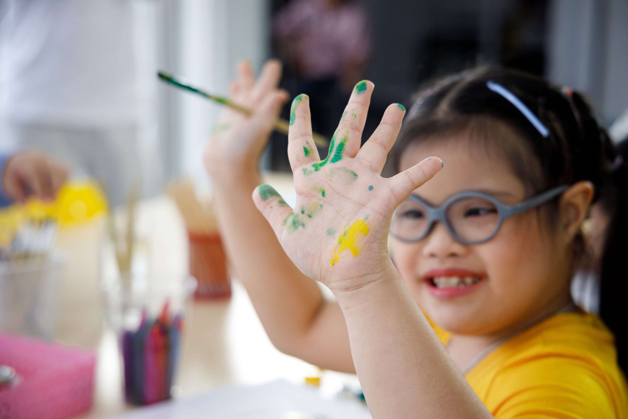 A child with additional support needs is painting. She is smiling and putting a paint covered hand up to the camera.