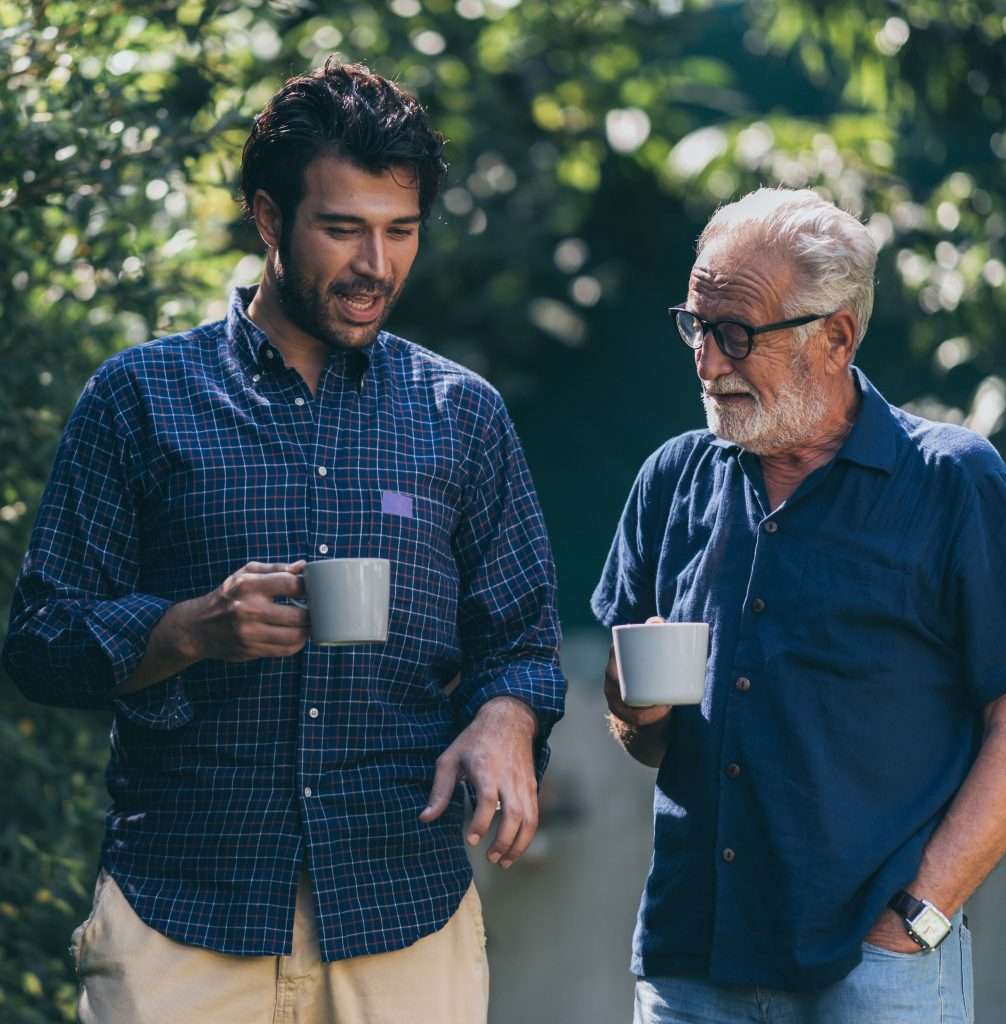 Two people outside having a chat. One is much older. They are holding mugs and smiling.