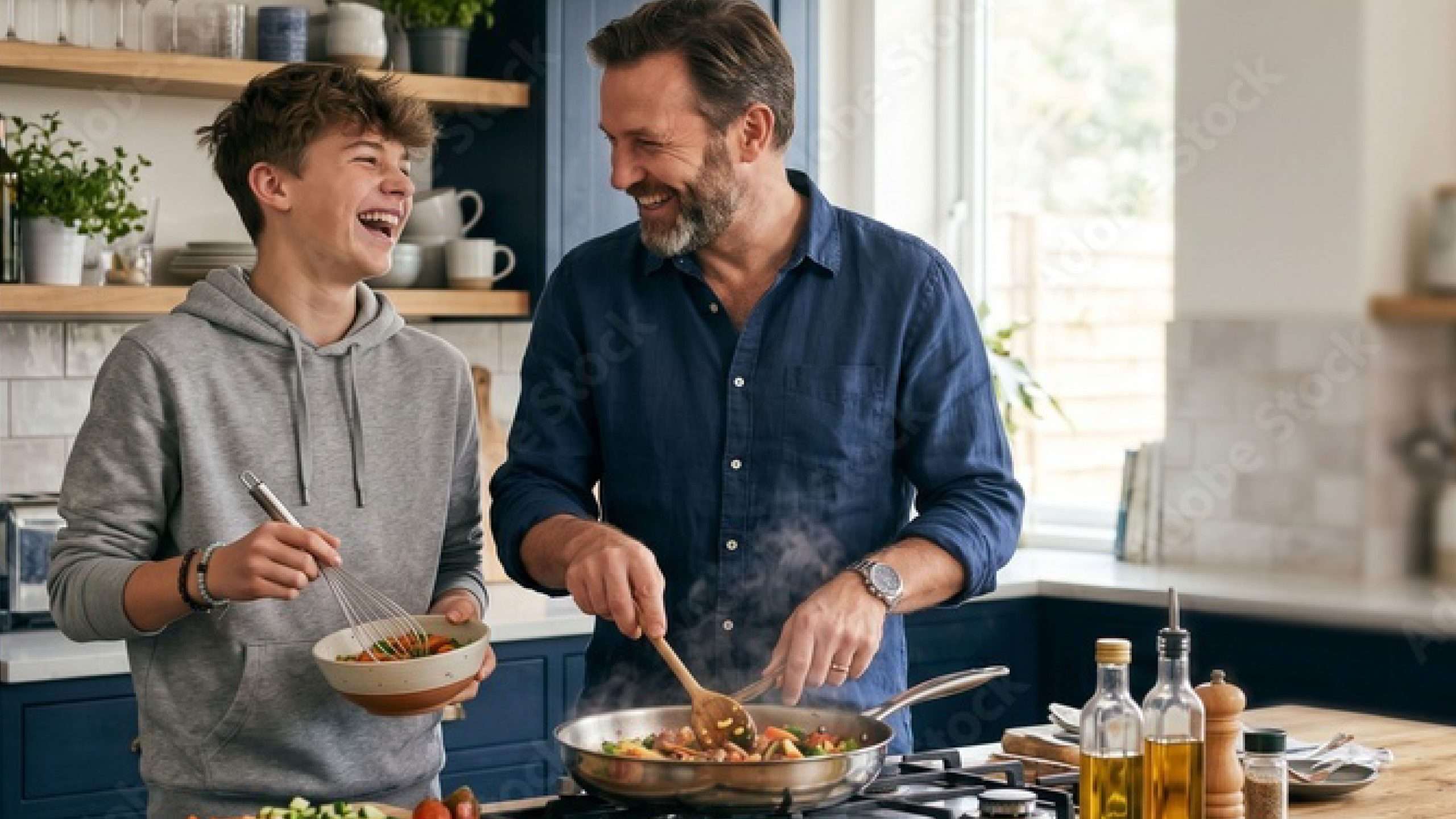 A young person is in the kitchen with an adult. They are cooking together and smiling. The young person has a bowl and the adult has a frying pan.